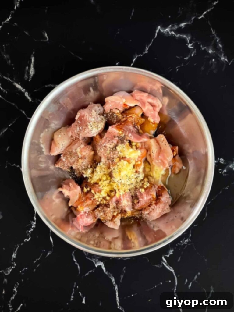 Diced chicken thigh pieces in a bowl with a mix of spices and marinade ingredients on top, ready to be combined.