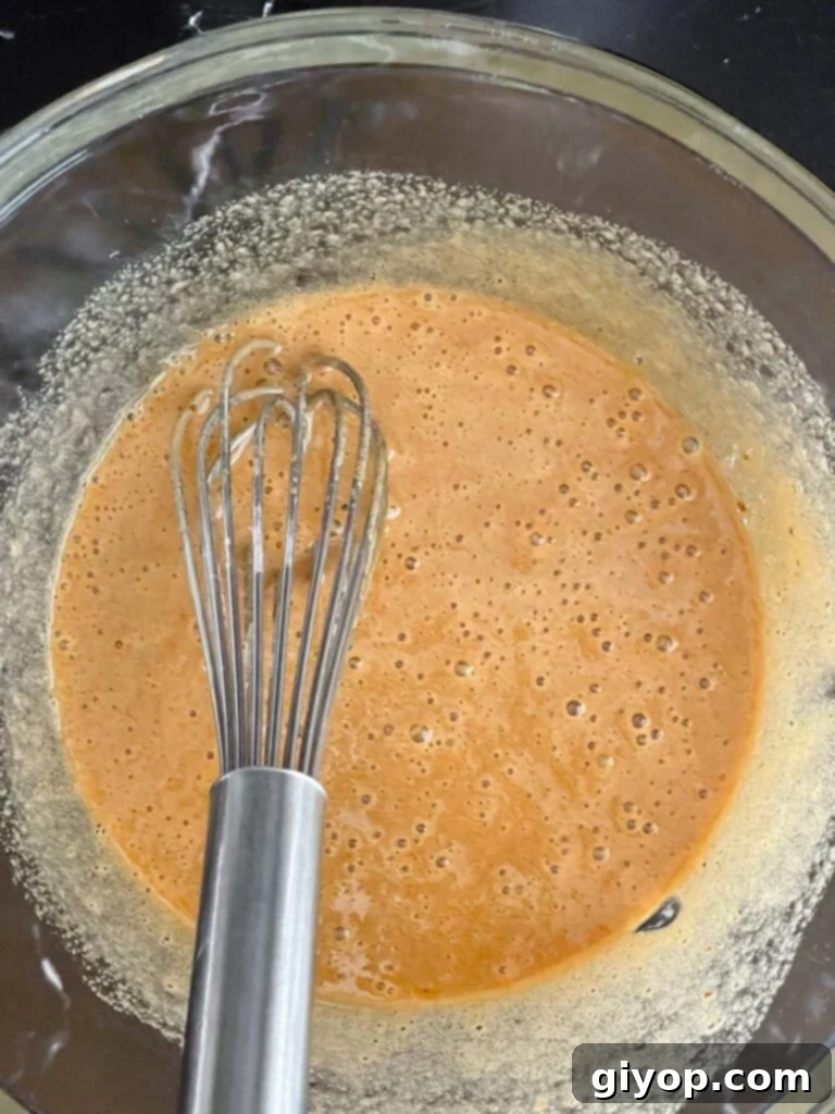 Wet ingredients, including whisked sugar and eggs, are mixed in a large glass bowl, creating a smooth and creamy mixture.