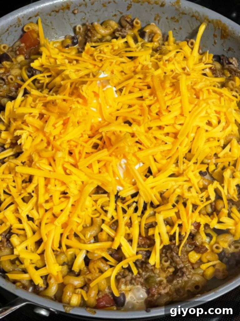 Shredded cheddar cheese being added to the hot taco pasta in a skillet, ready to melt into a creamy sauce.