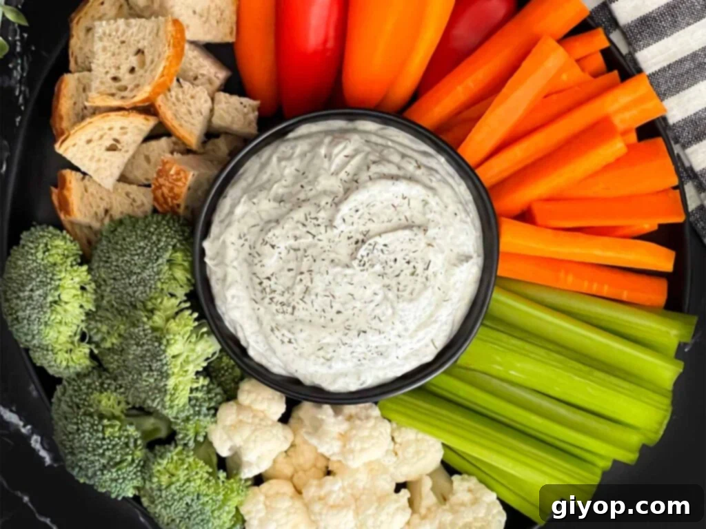 Dill dip in a small dark bowl surrounded by carrots, celery, cauliflower, broccoli, mini bell peppers and rye bread pieces on a dark plate.