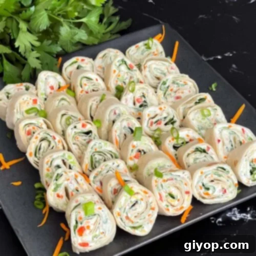 Veggie tortilla roll ups lined up on a square dark plate garnished with diced green onions and shredded carrots.