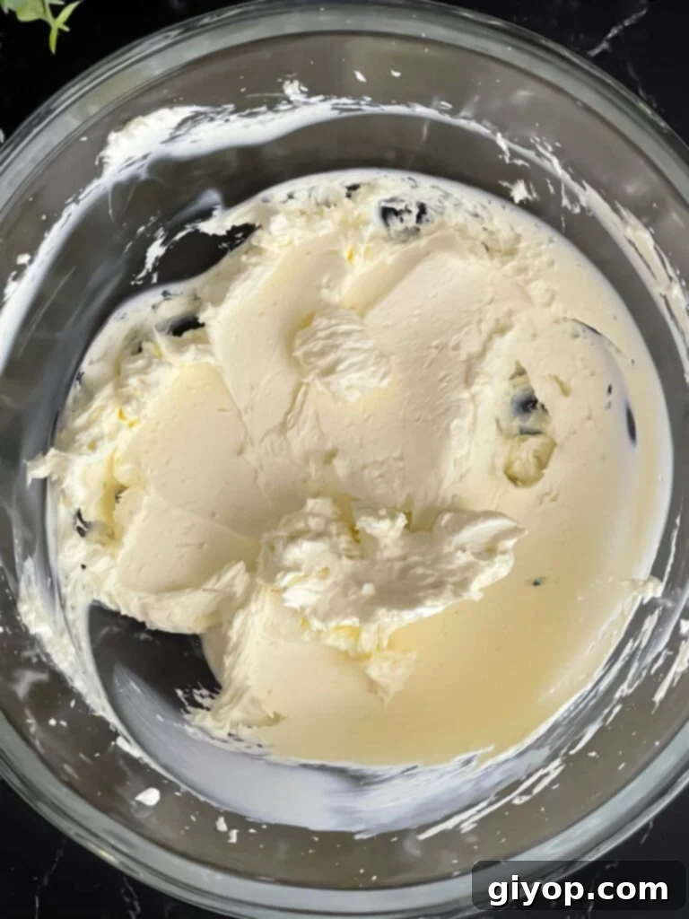 Softened cream cheese being beaten smooth in a large glass bowl on a dark surface, ready for the vegetable tortilla roll ups recipe.