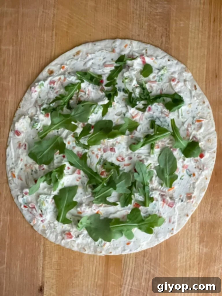 One large flour tortilla on a wooden cutting board, layered with the creamy vegetable mixture and fresh baby arugula, ready to be rolled.