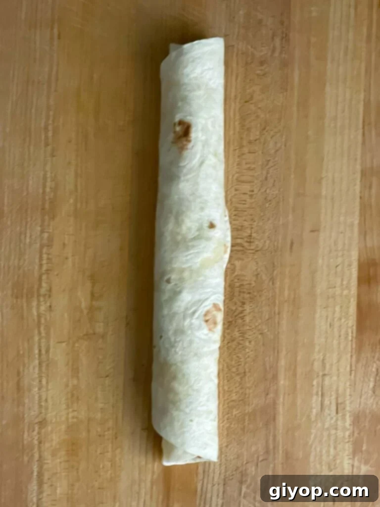 A tightly rolled veggie tortilla roll up on a wooden cutting board, ready for wrapping.