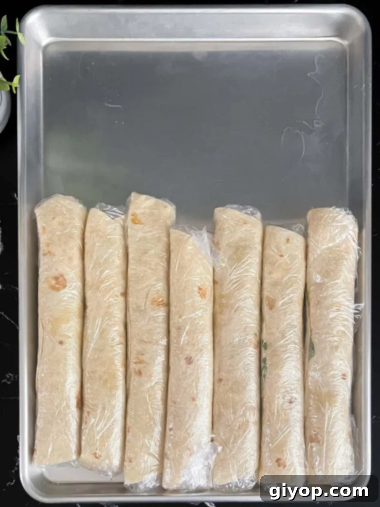 Several veggie tortilla roll ups, each individually wrapped tightly in plastic wrap, lined up on a baking sheet, ready for refrigeration.