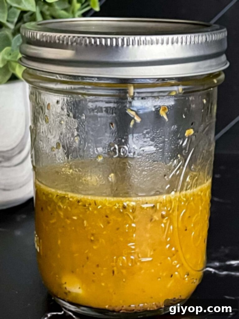 Zesty Greek Tortellini Pasta Salad 4 Homemade Greek dressing in a glass jar, showing separation of oil and vinegar before shaking, on a dark surface.