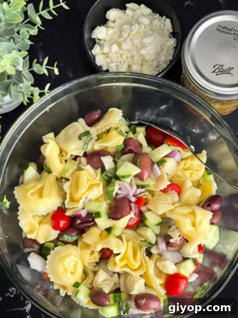 Zesty Greek Tortellini Pasta Salad 7 Cooked tortellini tossed with chopped vegetables in a large glass mixing bowl before the dressing is added.