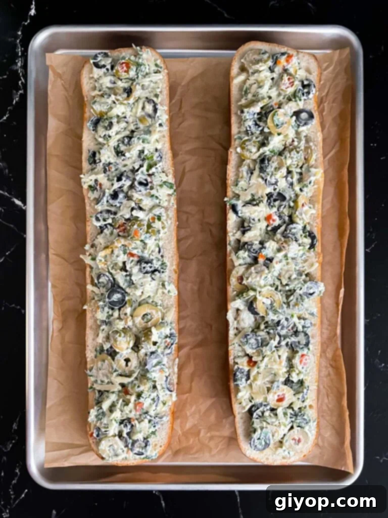 Unbaked olive cheese bread halves, generously topped with extra cheese, resting on a parchment paper lined baking sheet, ready for the oven.