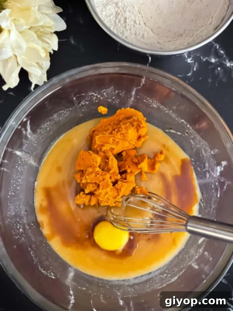 Blotted pumpkin puree, a large egg yolk, and vanilla extract being added to the wet ingredients in a glass mixing bowl.