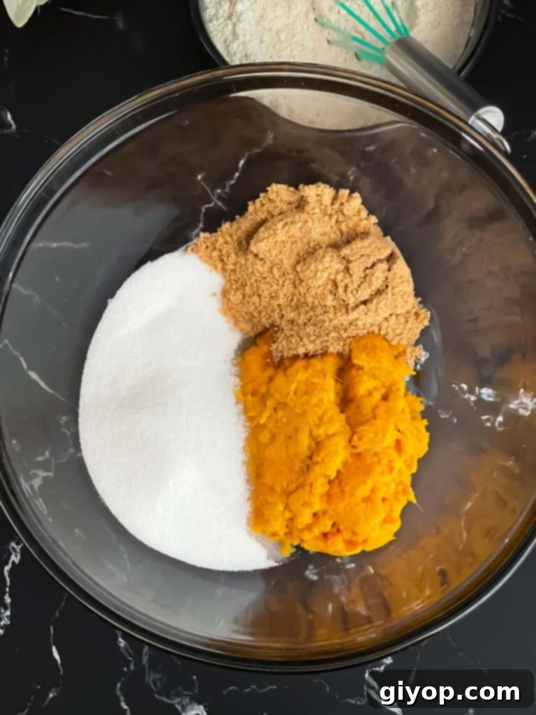 Sugars and mashed sweet potato in a glass mixing bowl on a dark surface, ready to be beaten.