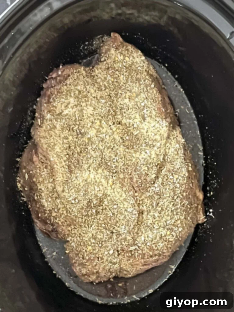 Chuck roast in a slow cooker, generously sprinkled with a mix of Italian seasonings.