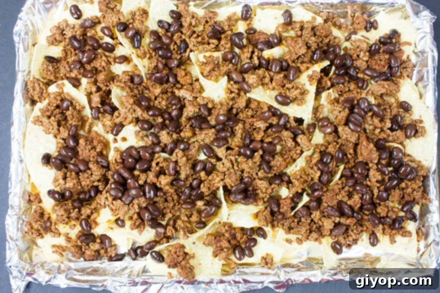 Loaded Sheet Pan Beef Nachos 4 Tortilla chips, seasoned ground beef, and black beans layered on a foil-lined baking sheet before baking.