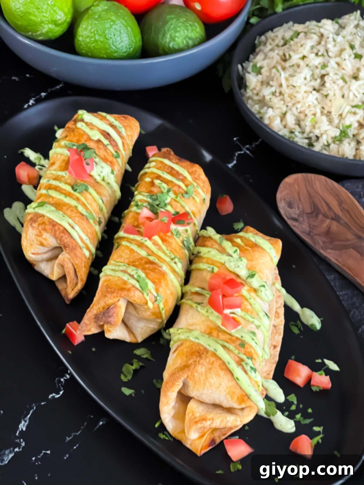 Baked chicken chimichangas on a dark oval platter garnished with avocado crema, diced tomatoes and cilantro.