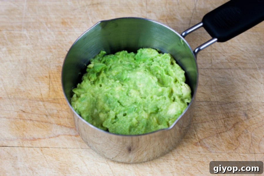 Creamy mashed avocado spread evenly over the cucumber layer in the metal measuring cup, forming the next level of the sushi stack.
