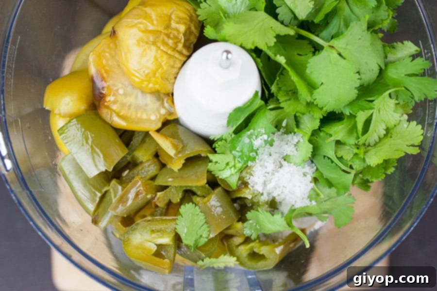 Roasted tomatillos, jalapeños, and onions placed in the bowl of a food processor, ready to be blended into homemade salsa verde.