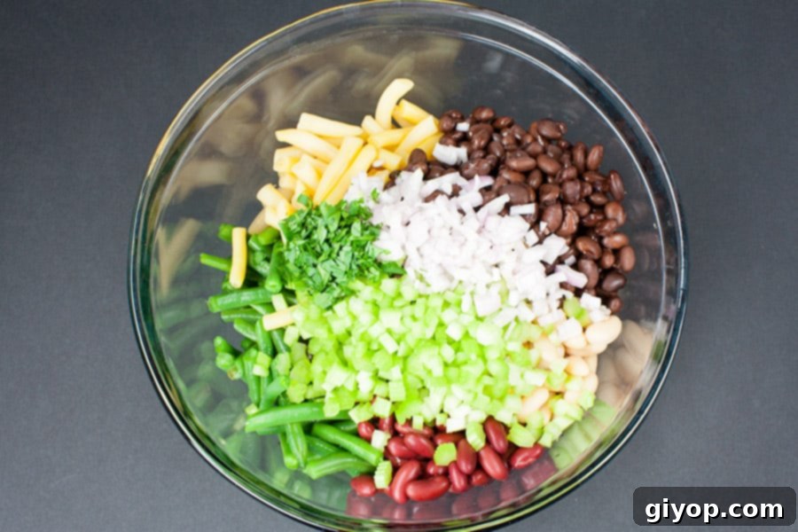 Ingredients for Five Bean Salad including various canned beans, fresh green beans, celery, shallots, and parsley, in a glass mixing bowl before mixing.