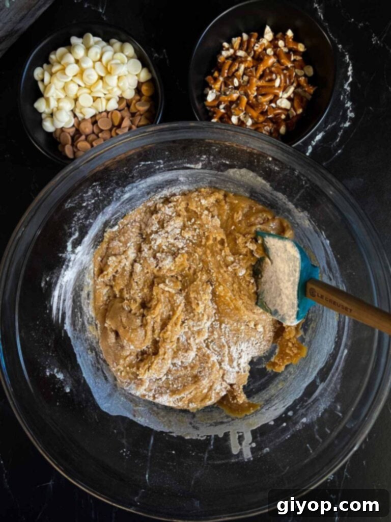 Mixing salted butterscotch pretzel cookie dough ingredients in a glass bowl on a dark surface.
