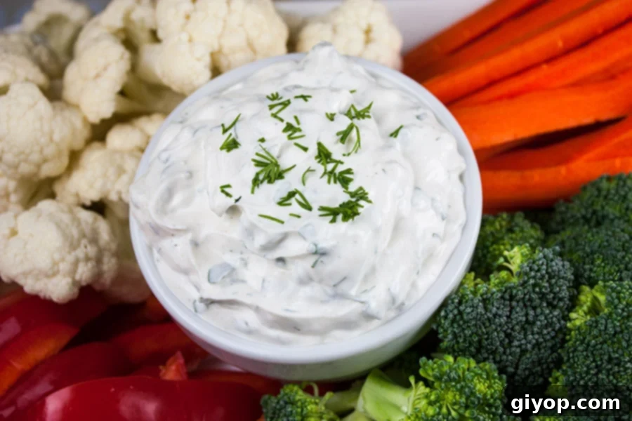A close-up of Fresh Herb Vegetable Dip in a pristine white bowl, garnished with a sprig of fresh dill, surrounded by an assortment of colorful cut vegetables including broccoli florets, cauliflower florets, carrot sticks, and vibrant red bell pepper slices, illustrating its perfect pairing.