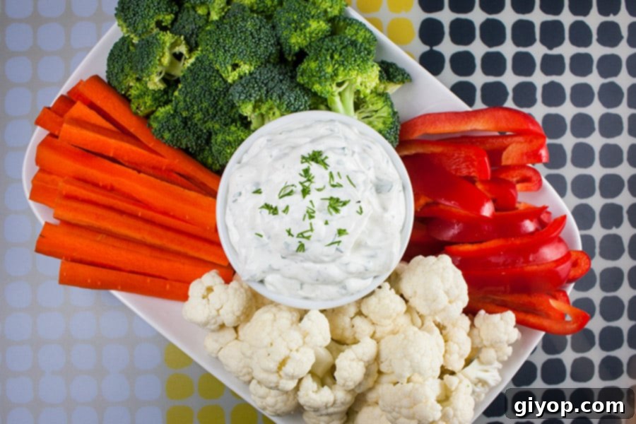 A pristine white bowl filled with Fresh Herb Vegetable Dip, beautifully garnished with a sprig of fresh parsley, nestled amongst an arrangement of colorful raw vegetables like broccoli, cauliflower, carrot sticks, and vibrant red bell pepper slices. The image highlights the freshness and appeal of the dip, ready to be enjoyed.